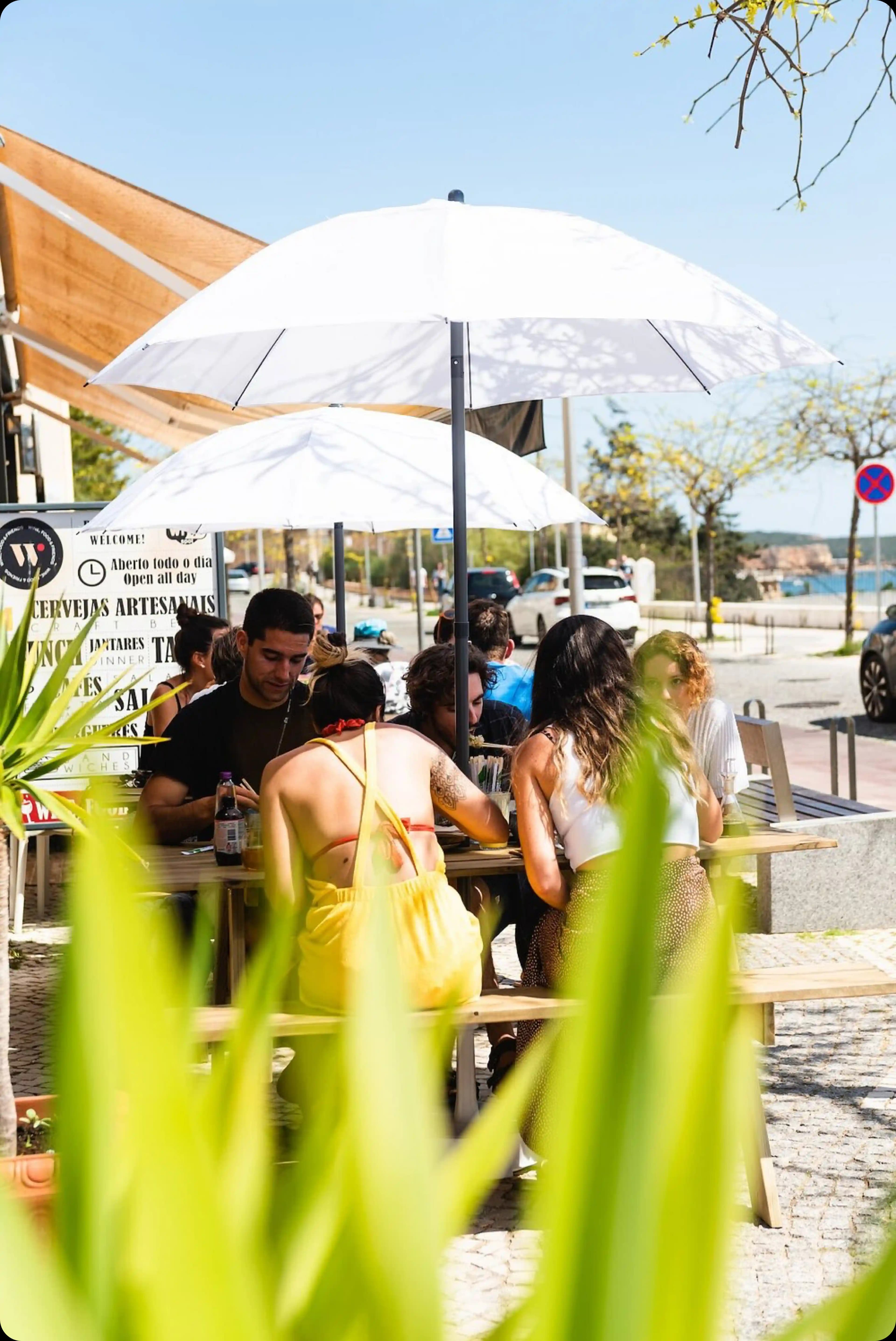 A group of people enjoying food and conversation on the sunny outdoor patio under a large white umbrella.