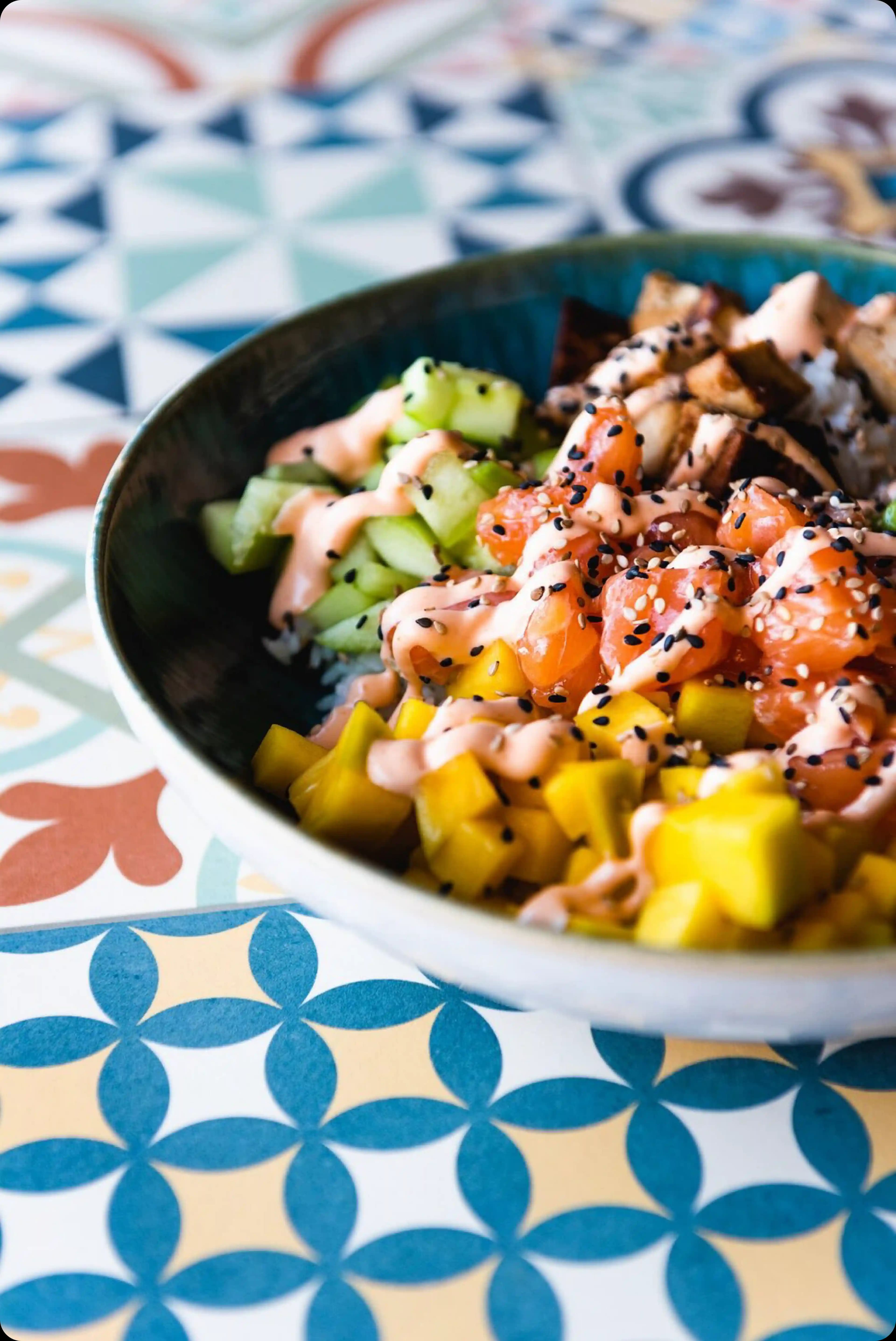 A fresh poké bowl with salmon and mango served on a table with blue and white geometric tiles.