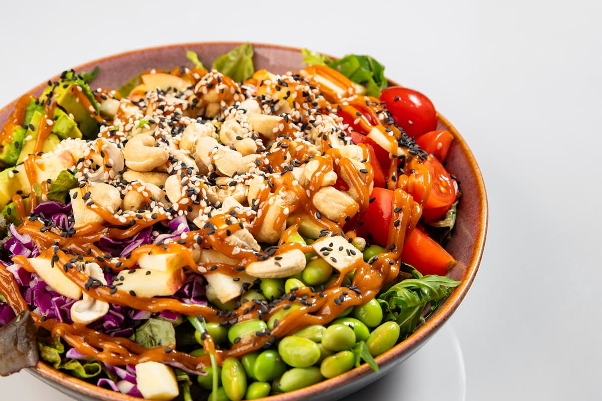 A vibrant, healthy poke bowl with cashews, tomatoes, salad and edamame, topped with some sauce and sesame seeds.
