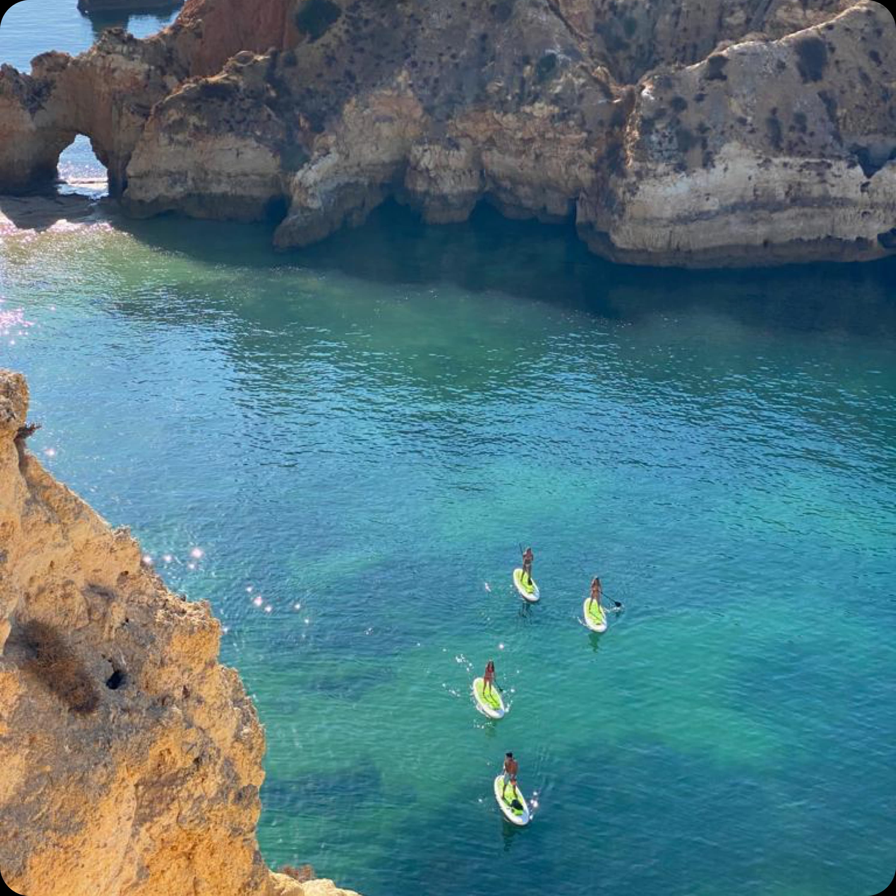 An aerial view of paddleboarders in a beautiful turquoise water cove surrounded by golden cliffs in the Algarve.